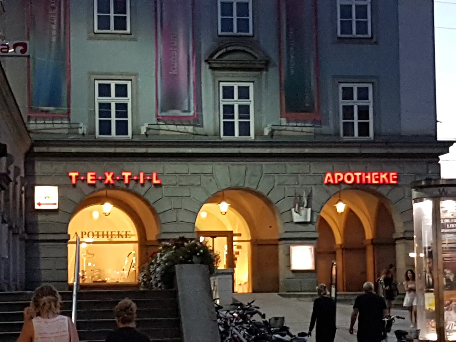 Beleuchtete Arkadenbögen des Brückenkopfgebäudes am Abend mit rotem Leuchtschriftzug „Apotheke“