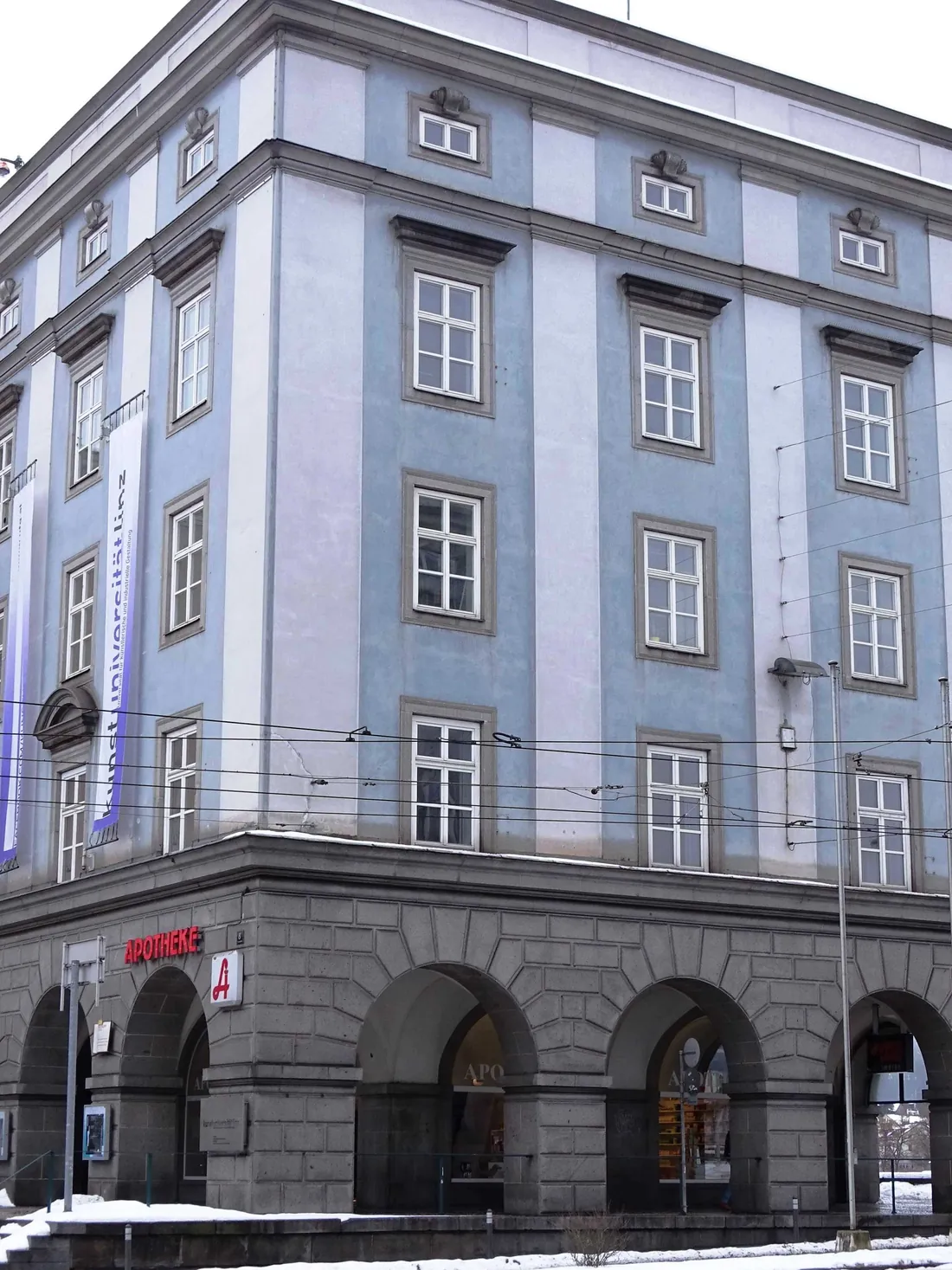 Hellblaue Fassade des Brückenkopfgebäudes West mit Arkaden im Erdgeschoß und rotem Apotheken-A