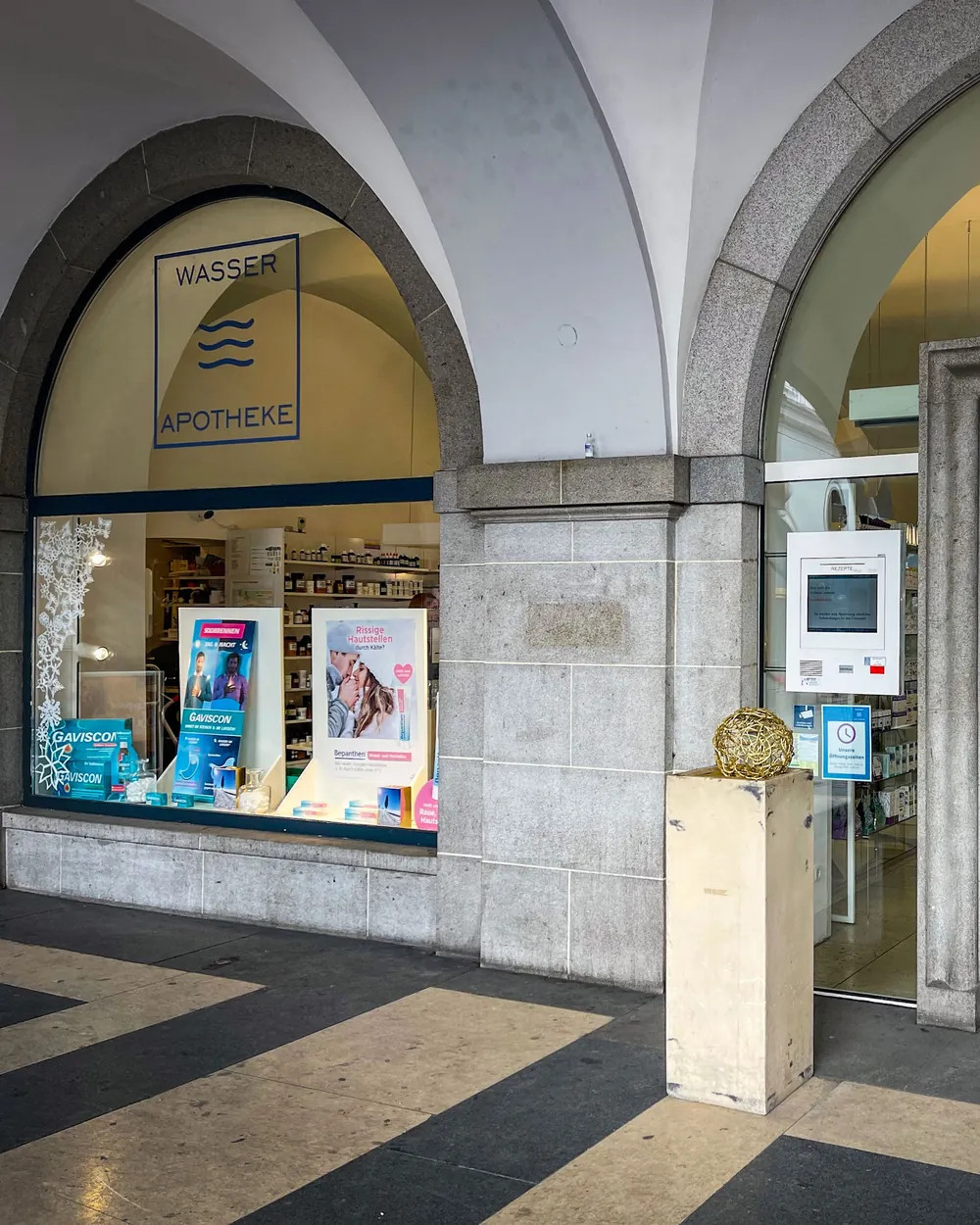 Glastür und Rundbogenfenster der Wasser-Apotheke mit dem Wellen-Logo unter den Arkaden des Brückenkopfgebäudes