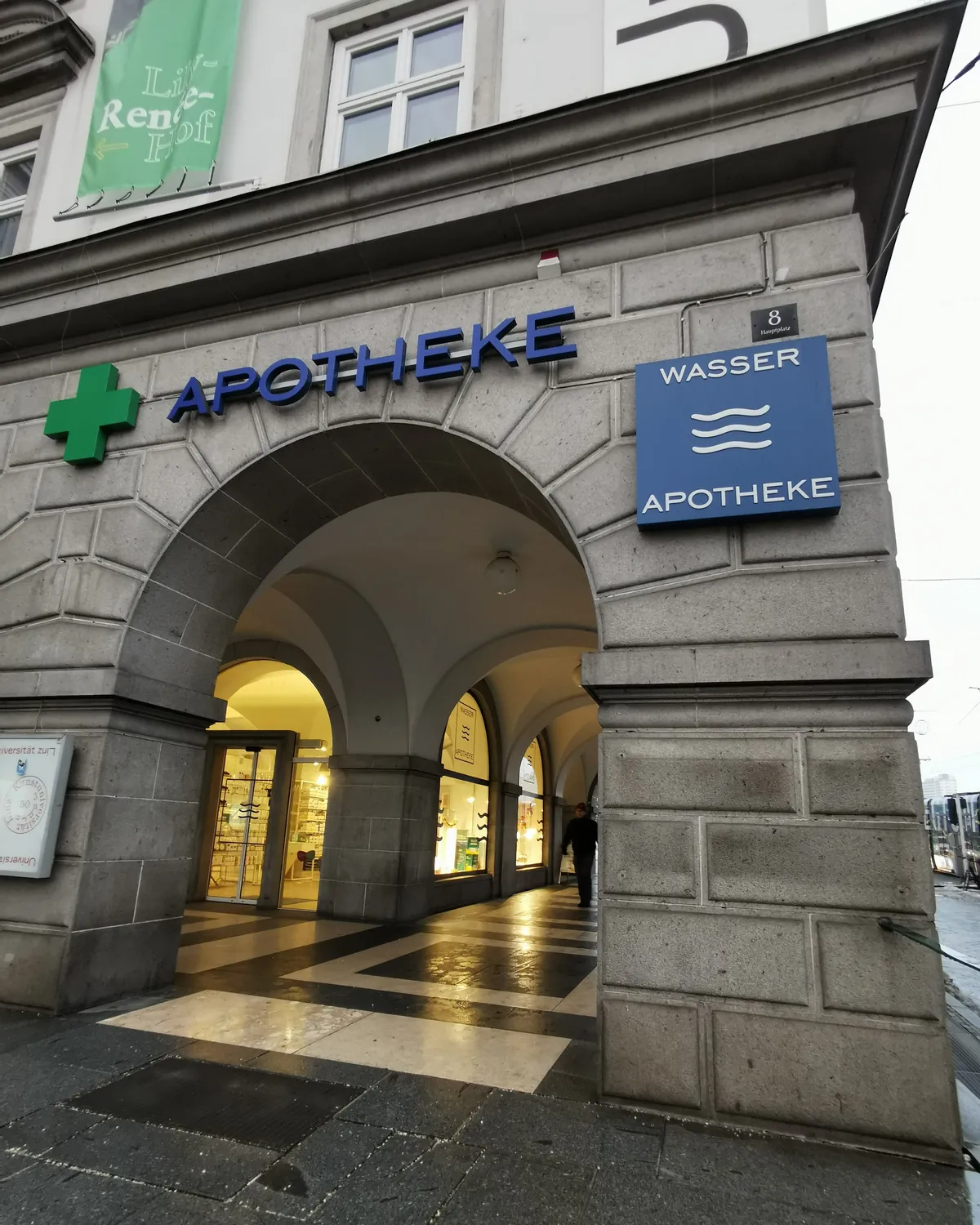 Steinerner Arkadenbogen mit grünem Apothekenkreuz, blauem Schriftzug „Apotheke“ und dem Schild „Wasser Apotheke“, dahinter der beleuchtete Eingang