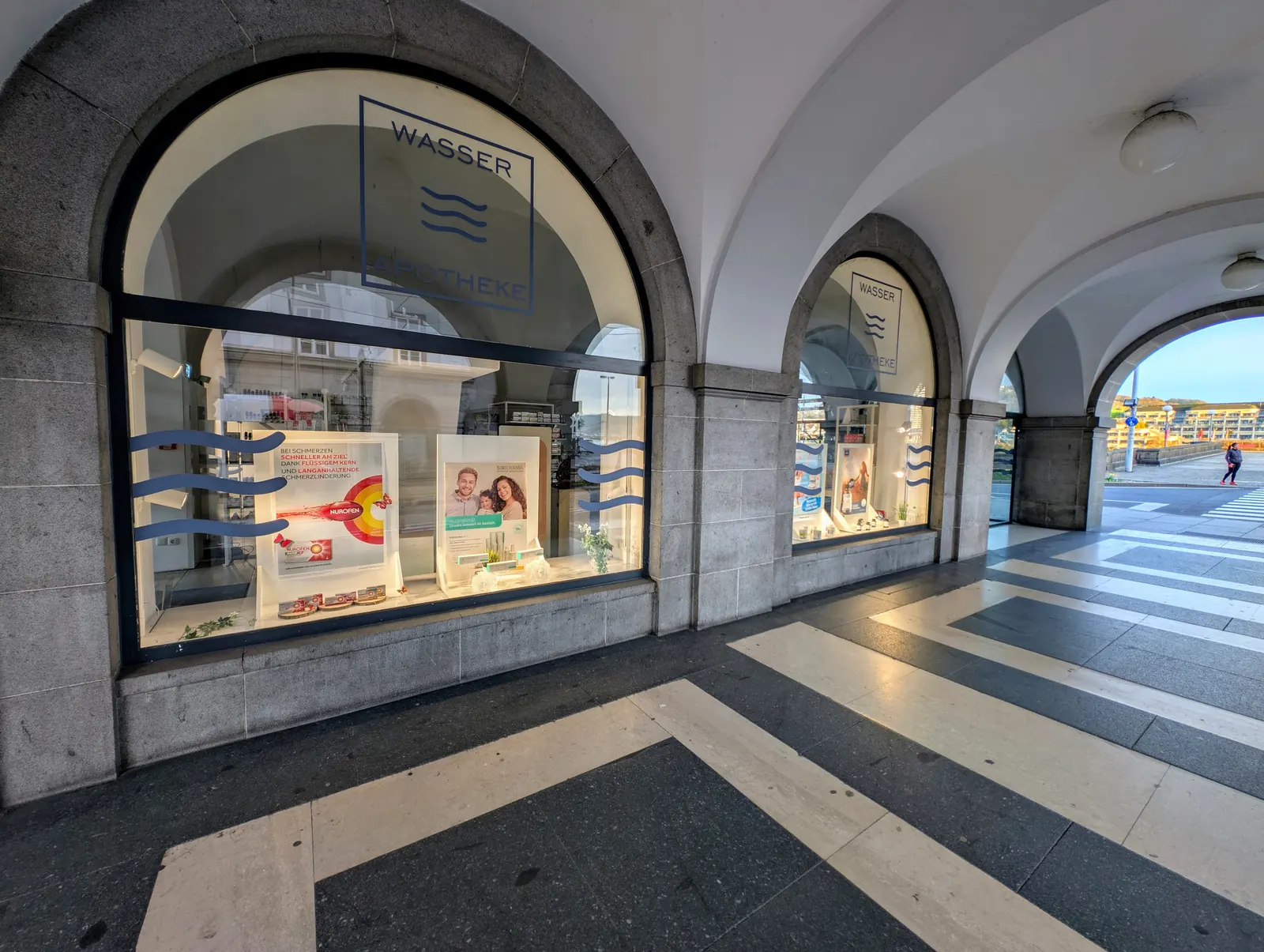 Rundbogen-Schaufenster der Wasser-Apotheke mit Wellen-Logo im Arkadengang, hinten der Blick auf die Donau
